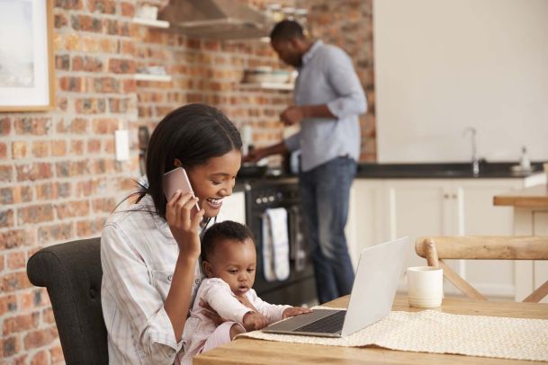 Woman and baby at table on phone in front of laptop