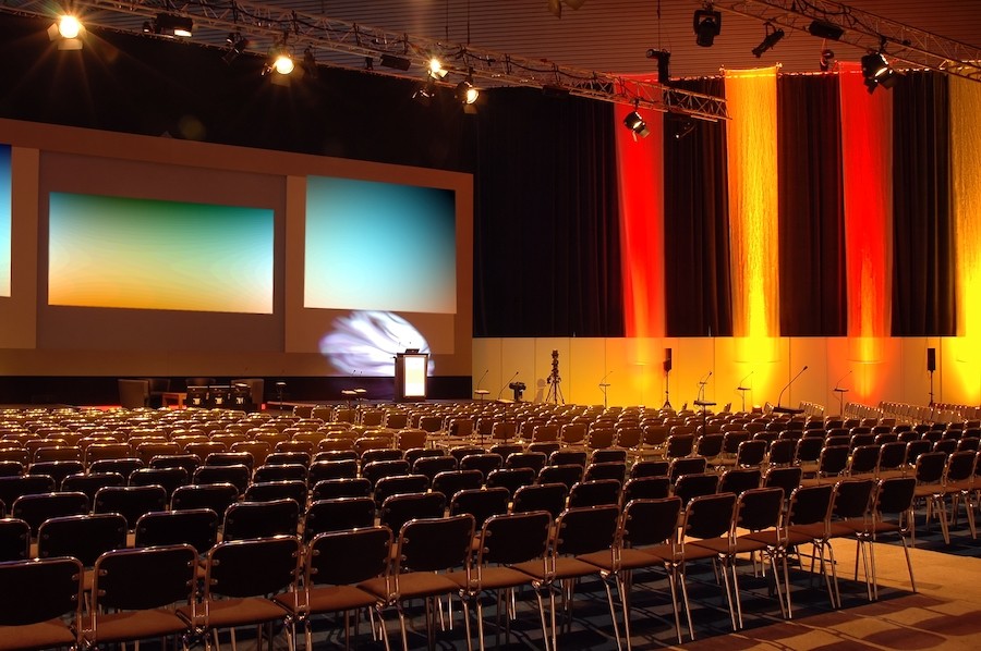 Large conference space with rows of chairs, red and orange lighting, and three large screens on a stage.
