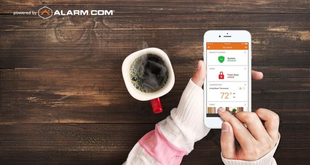 Woman holding phone with alarm.com interface on it next to coffee
